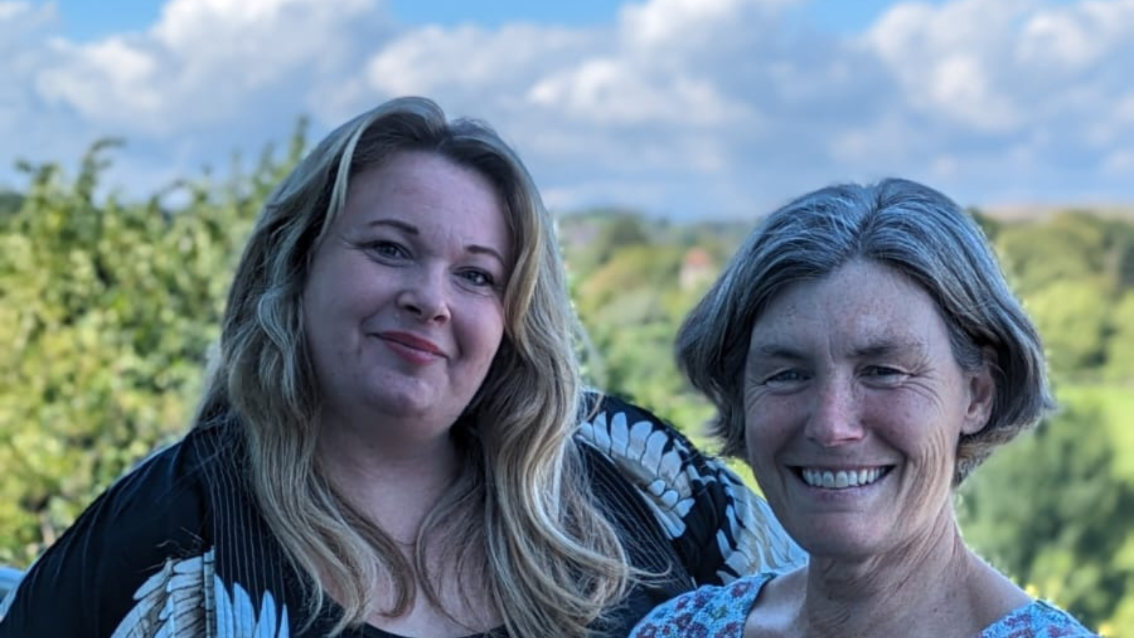 Photo of two white women outside with trees in the background. stood next to each other and smiling towards the camera. The woman on the left has long hair and the woman on the right has a short bobbed hair cut.