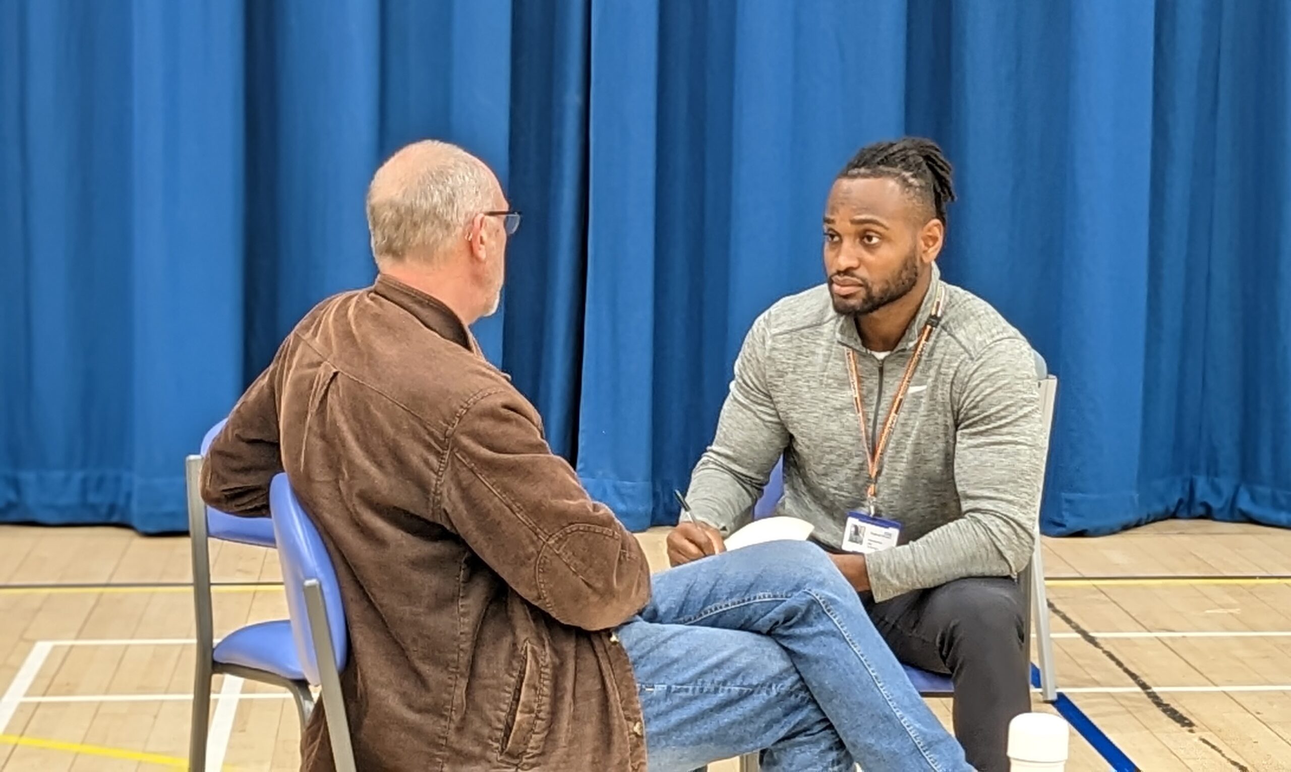 An older man wearing a jacket and jeans is sat with his legs crossed talking to a younger black male clinician - who is listening and taking notes at a Community Appointment Day