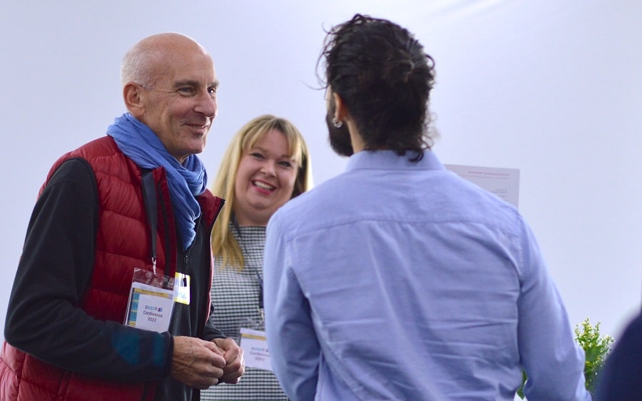 Photo of two white women outside with trees in the background. stood next to each other and smiling towards the camera. The woman on the left has long hair and the woman on the right has a short bobbed hair cut.