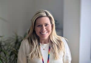 A Rethinking Our Health workshop space featuring flipcharts and whiteboards covered in colourful sticky notes. One flipchart is labelled "SOLUTIONS" with a hand-drawn lightbulb in the centre, surrounded by ideas on post-it notes. In the background, posters titled "YOUR HEALTH" and "BARRIERS" are visible, with more sticky notes and diagrams. The image is framed by an abstract teal shape.