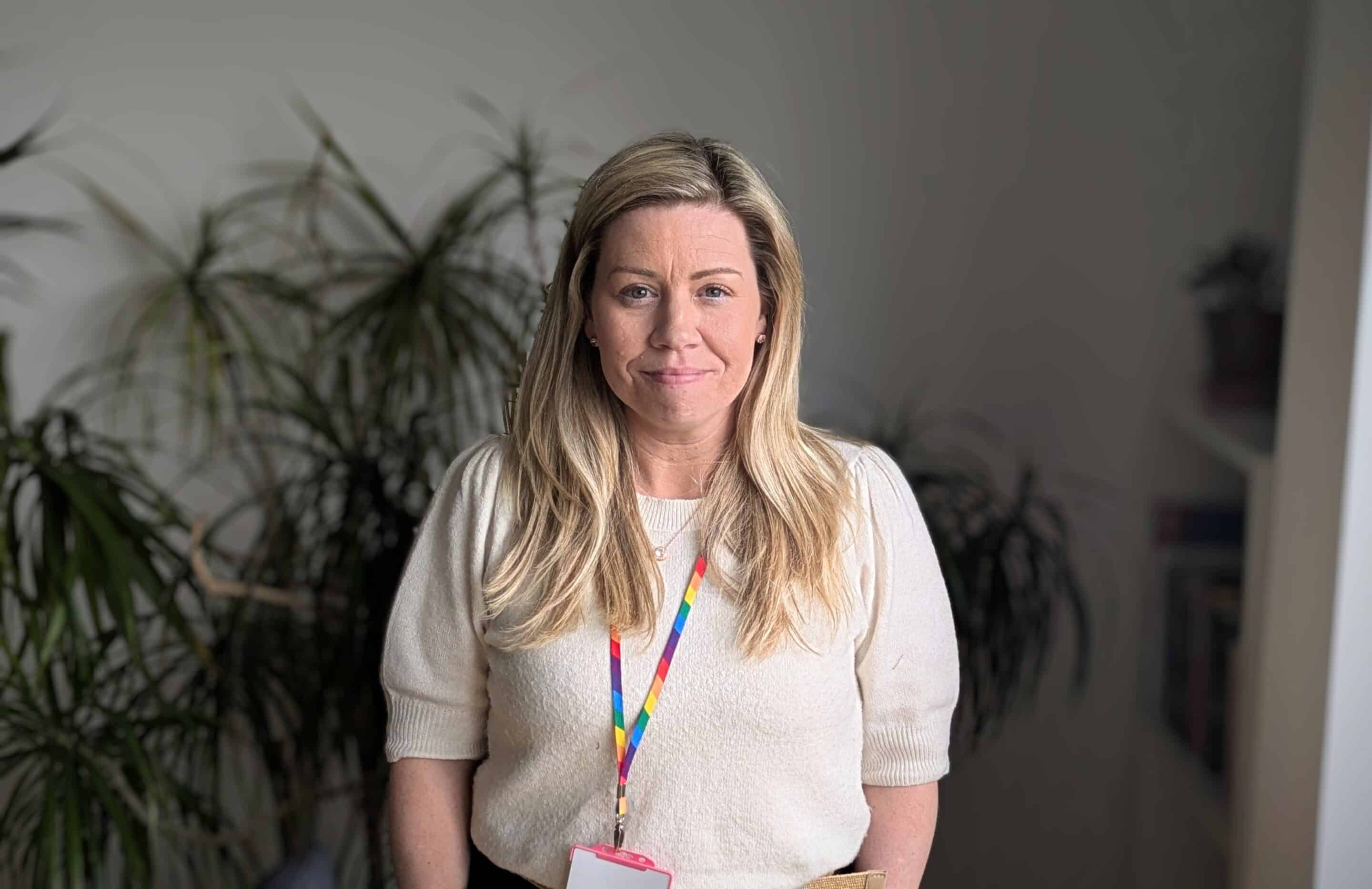 Photo of two white women outside with trees in the background. stood next to each other and smiling towards the camera. The woman on the left has long hair and the woman on the right has a short bobbed hair cut.