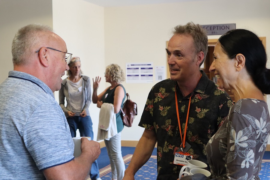 Photo of two white women outside with trees in the background. stood next to each other and smiling towards the camera. The woman on the left has long hair and the woman on the right has a short bobbed hair cut.