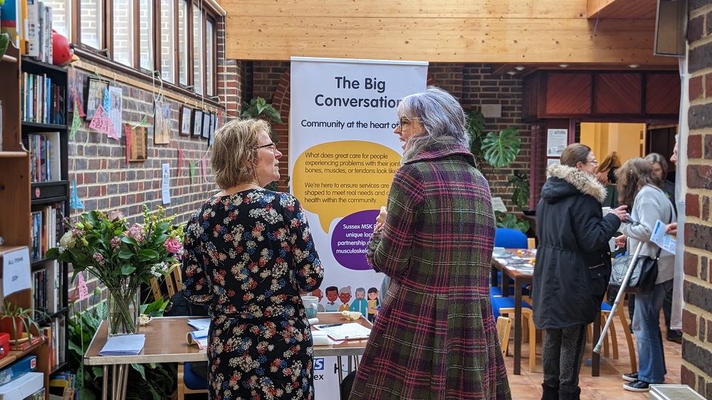 Photo of two white women outside with trees in the background. stood next to each other and smiling towards the camera. The woman on the left has long hair and the woman on the right has a short bobbed hair cut.