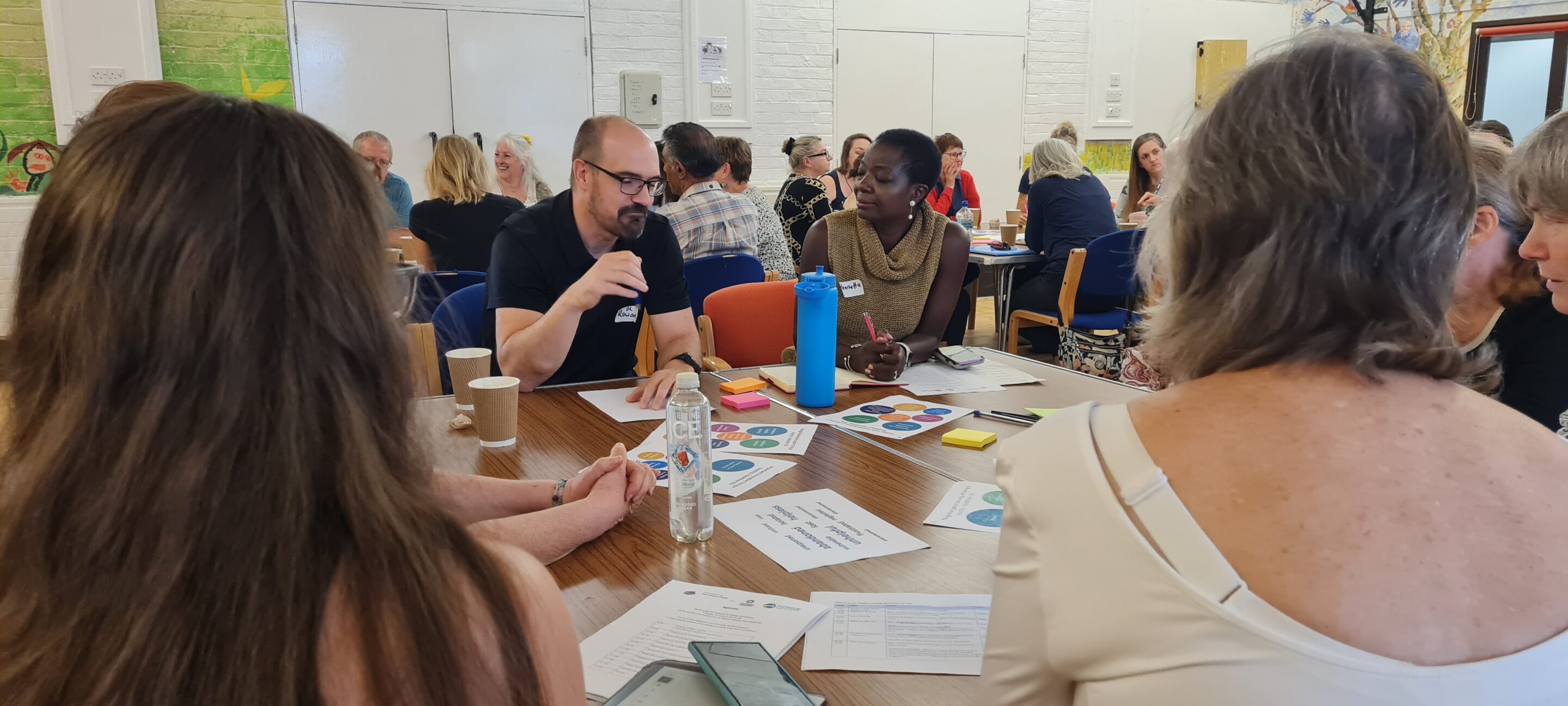 A group of people participating in a roundtable discussion during a community codesign event. The table is covered with printed worksheets, post-it notes, and drinks. The setting is a bright room with murals on the walls and multiple groups in conversation