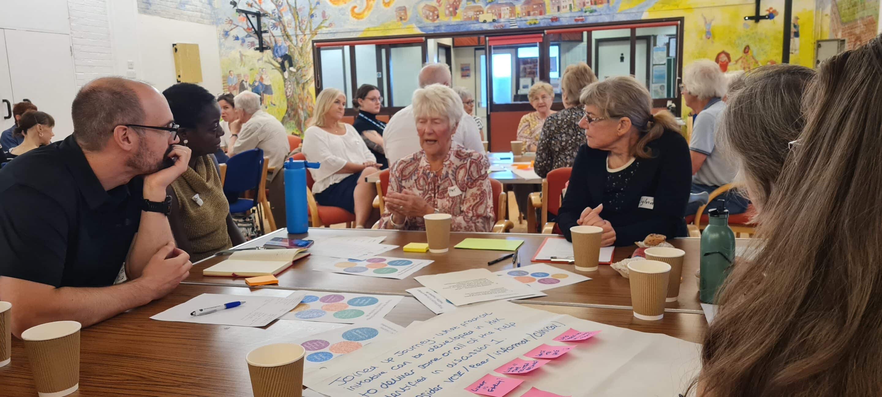 A group of people of various ages sit around a table in a bright, community-style room, engaged in lively discussion. Papers, pens, coffee cups, and post-it notes are spread out on the table. A colourful mural and more participants are visible in the background, contributing to a collaborative and inclusive atmosphere
